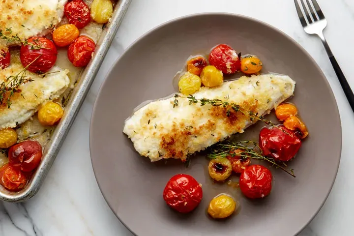 breaded fish fillet and tomatoes on gray plate with tray and fork on marble background